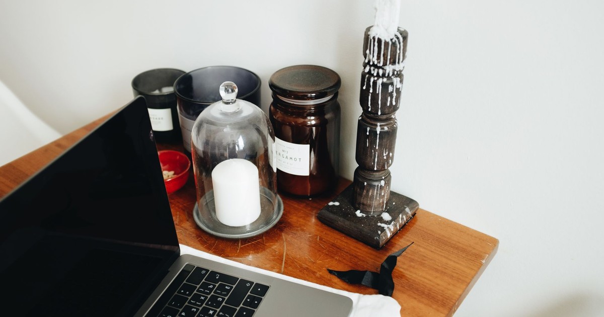 Freelance workspace with laptop and various black aromatic candles in glass jars and wooden candlestick placed on wooden table in light room of modern apartment