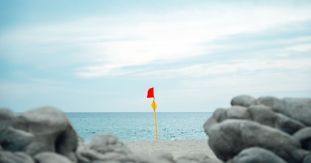 A red warning flag on a sandy beach in Oaxaca, Mexico, with ocean and rocky foreground.