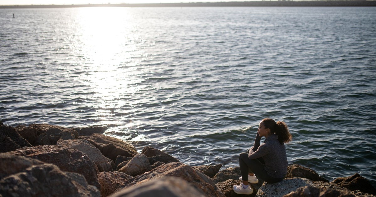 A woman in a sweater sits on rocks by the sea under bright daylight.