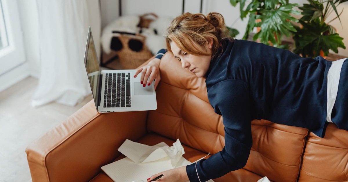Woman resting on a couch with laptop, surrounded by papers, depicting stressed remote work lifestyle.