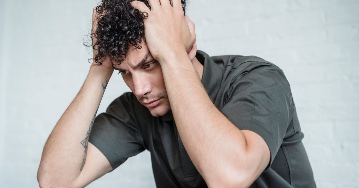 A young man with curly hair looks stressed as he sits with his hands on his head in contemplation.
