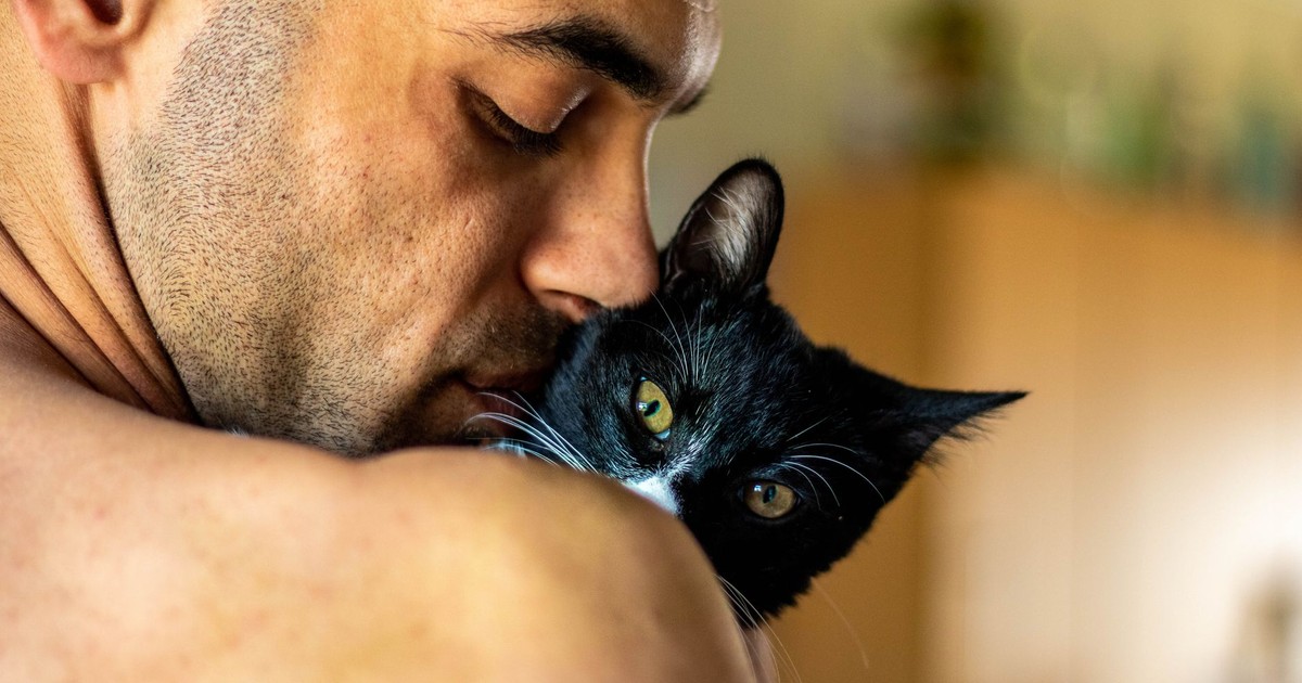 A close-up of a man affectionately holding a black and white cat in a warm home setting.