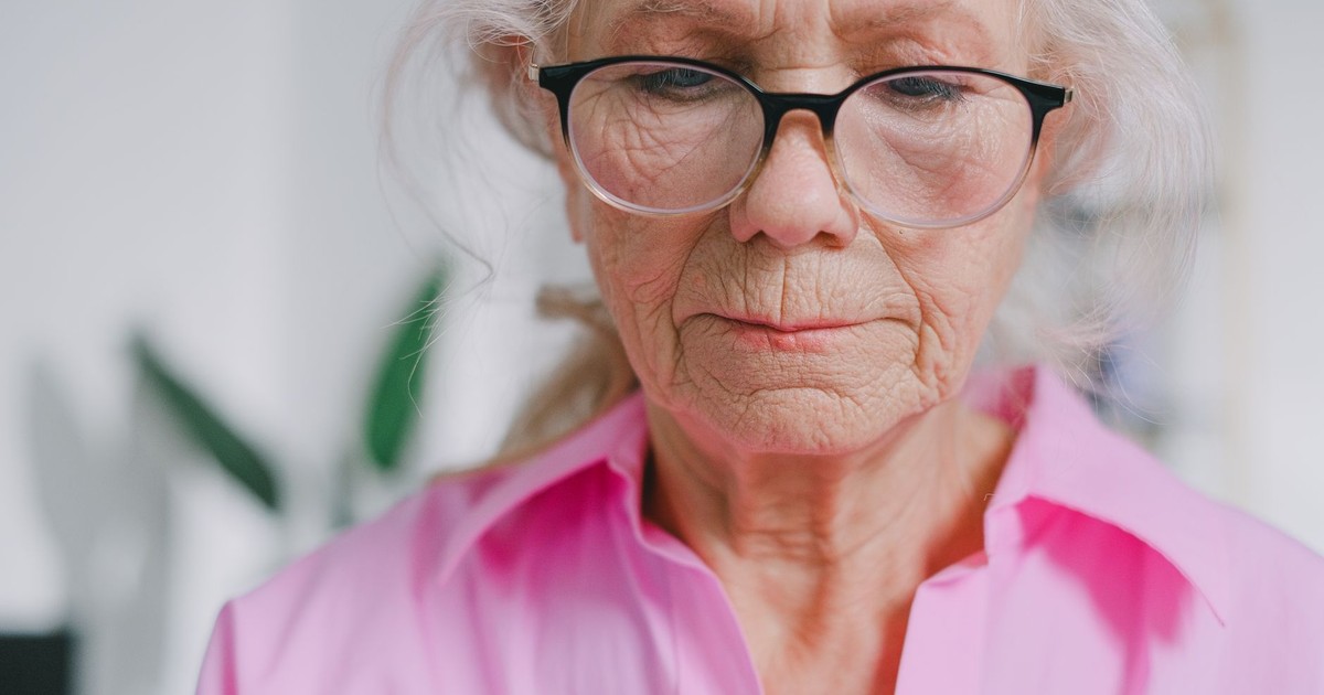 Portrait of an elderly woman in a pink shirt, focusing thoughtfully indoors.