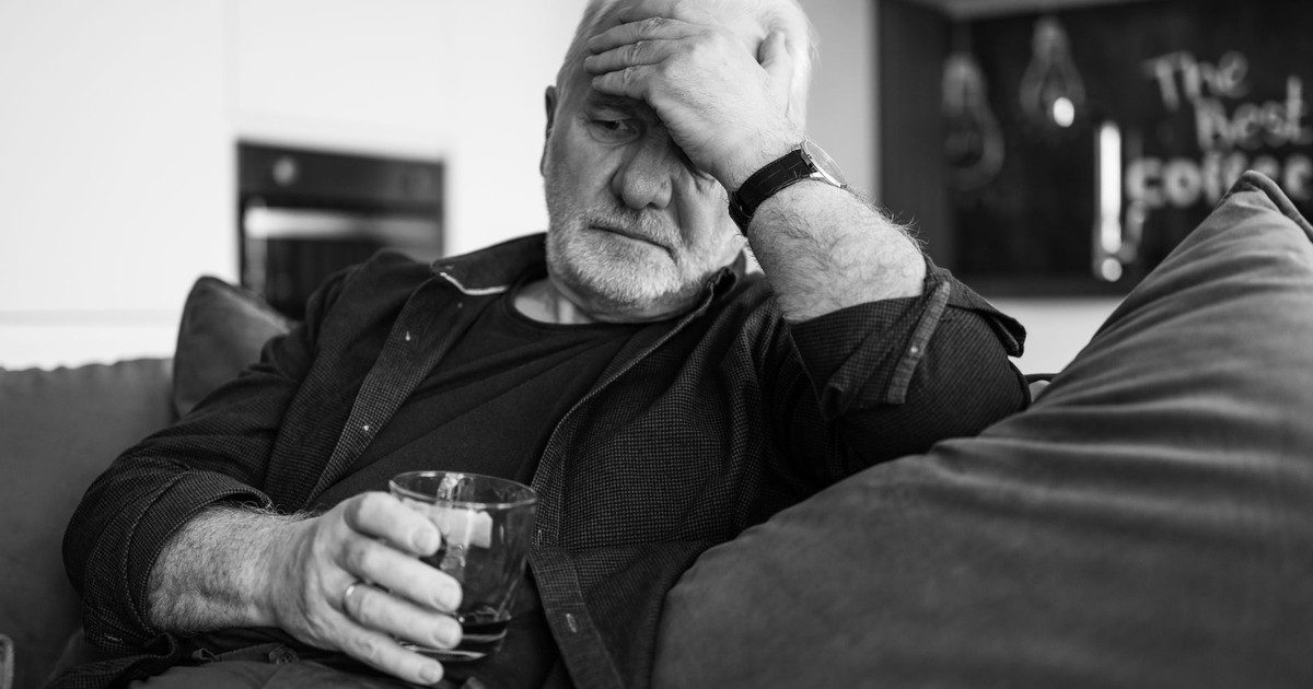 Grayscale image of an elderly man sitting indoors, holding a drink with a hand on his forehead, conveying emotion.