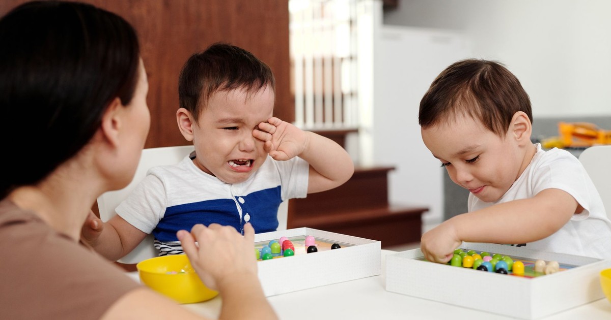 A mother attending to her crying child while another child plays contentedly indoors.