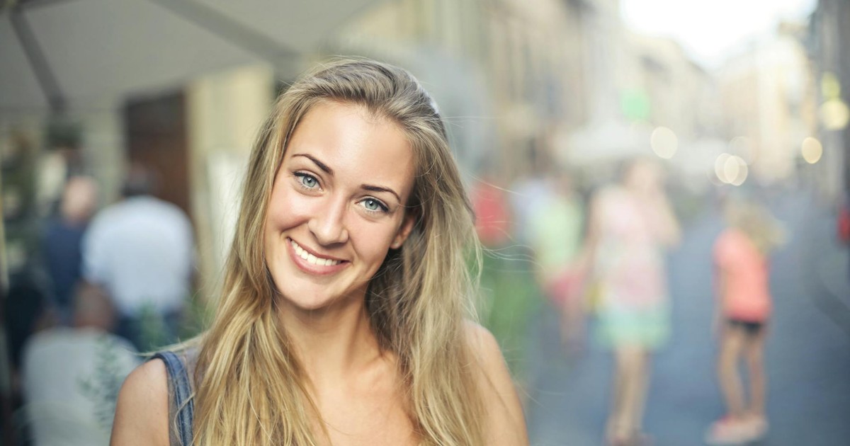 Portrait of a smiling woman with blonde hair standing outdoors in a bustling street setting.