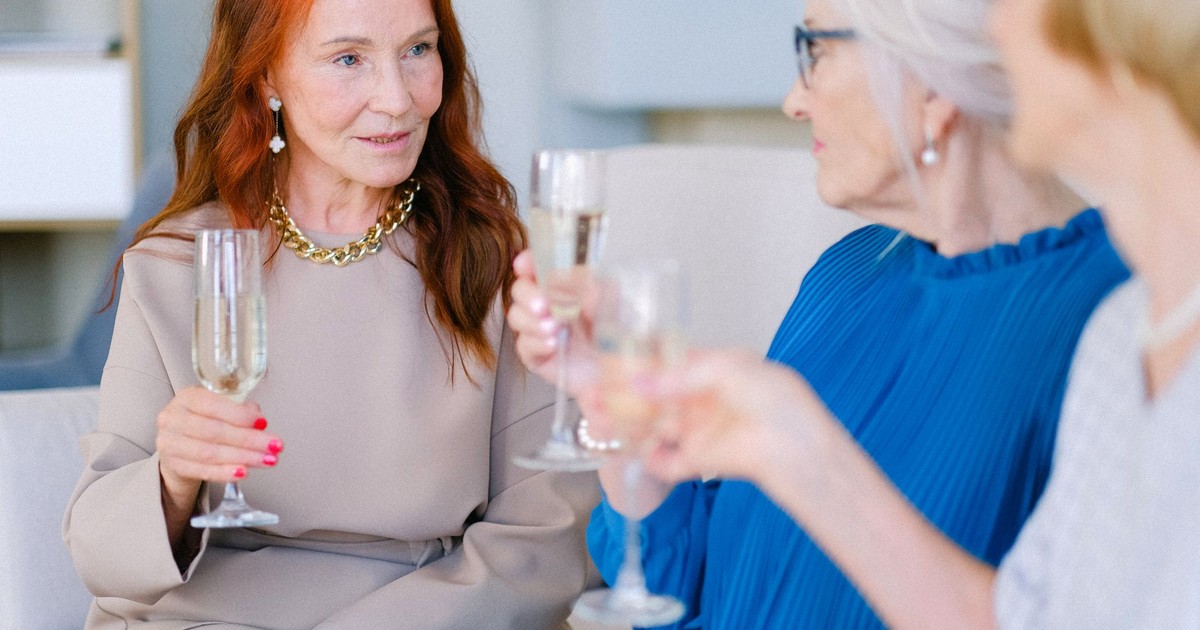 Group of aged crop women making toast with glasses of champagne on blurred background of living room