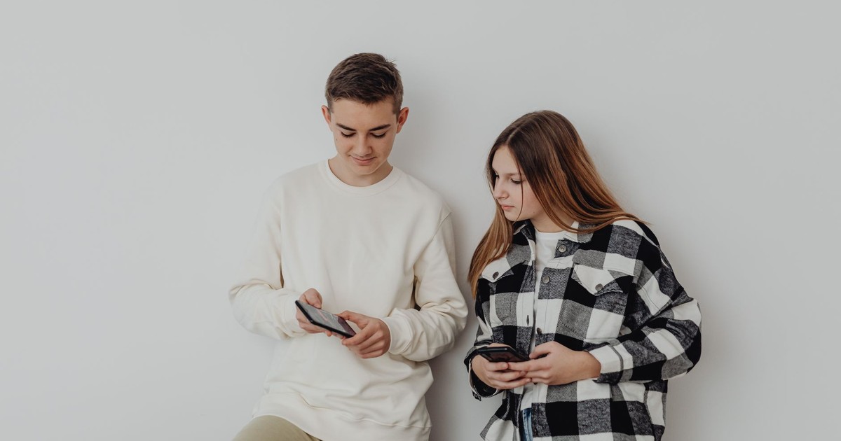 Teenage boy and girl using smartphones while leaning against a white wall indoors.