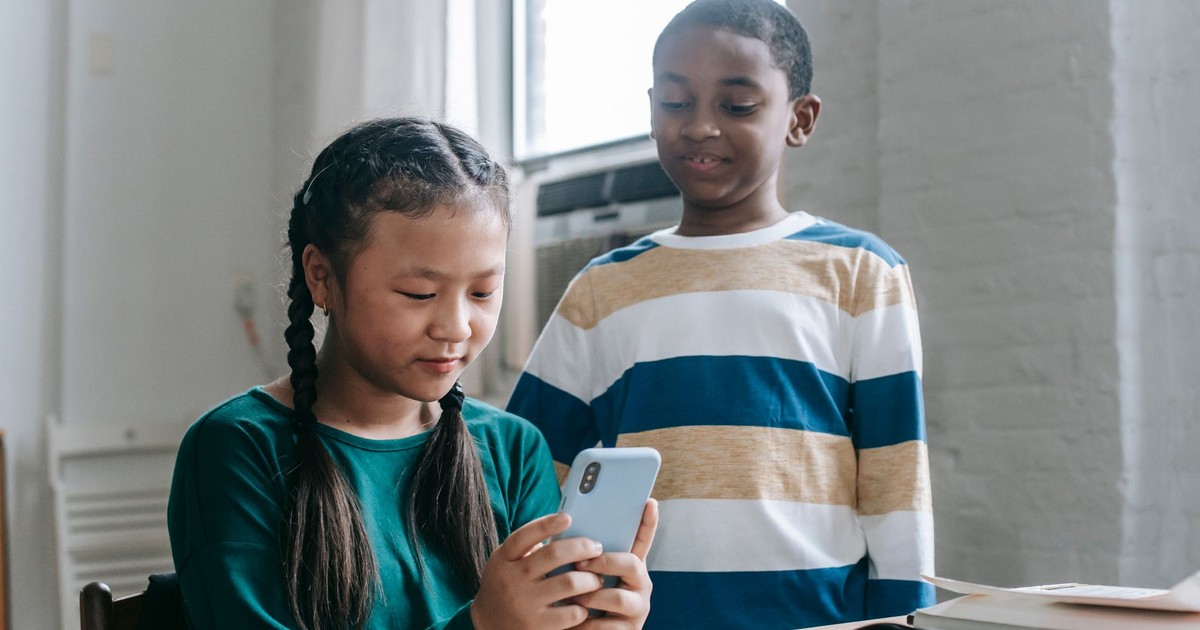 Happy little African American boy standing near Asian classmate girl using smartphone sitting at desk during break