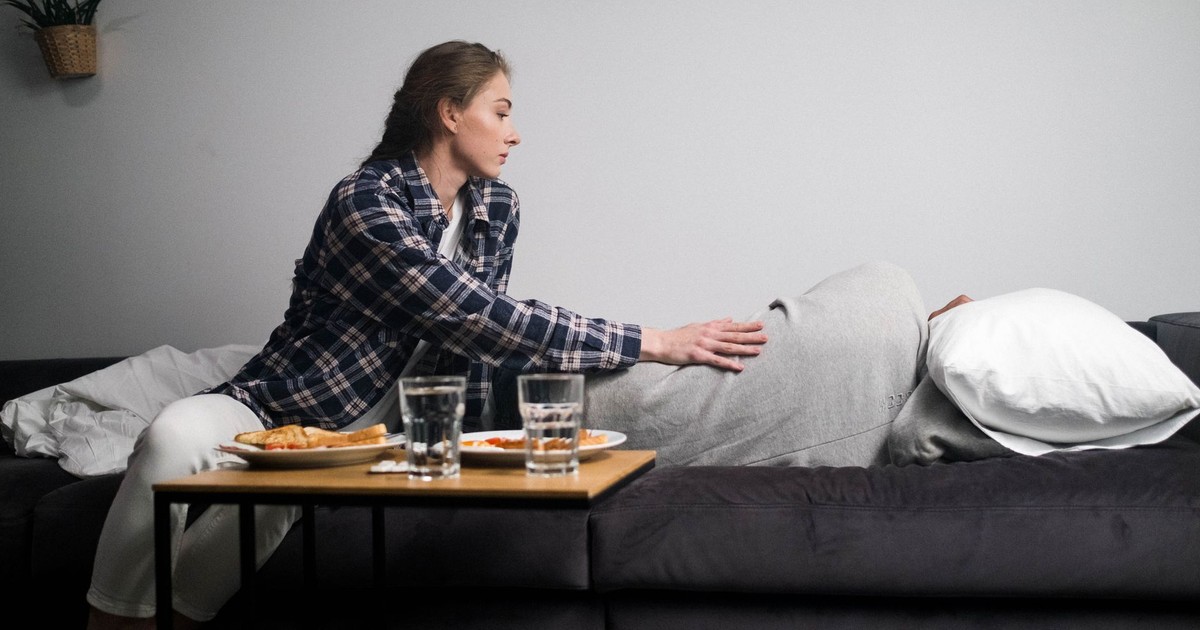 A woman comforts her partner lying on a couch beside a breakfast tray.