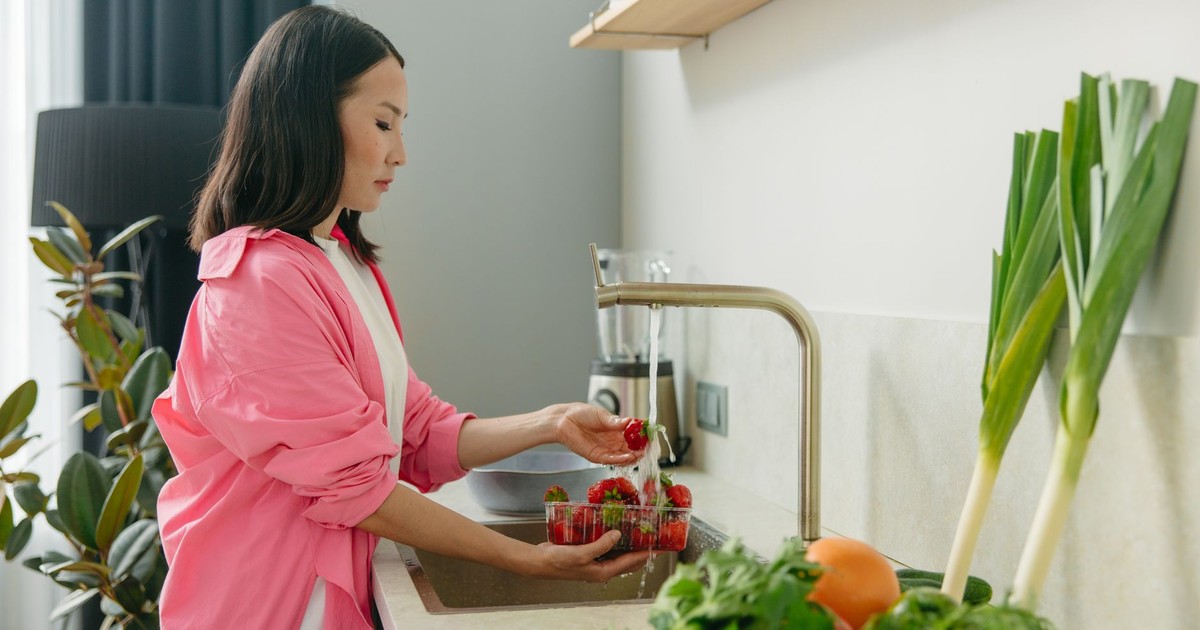 Asian woman washes strawberries at kitchen sink, promoting healthy living.