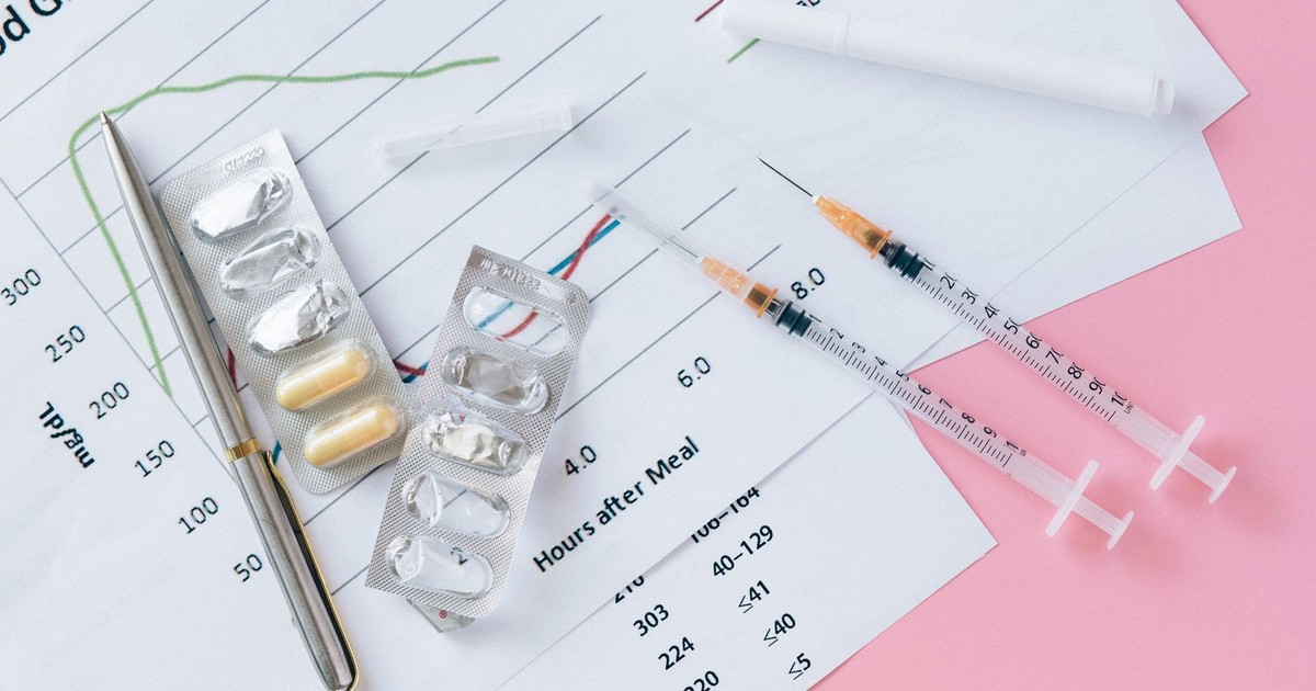 Flat lay of medicines, syringes, and a blood glucose chart on a pink surface, representing health management.