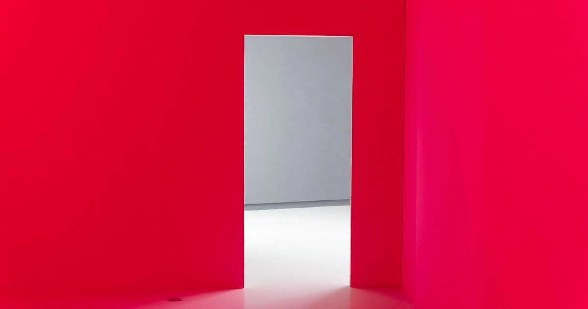 Abstract interior with bright red walls and an open doorway leading to a minimalist white room.