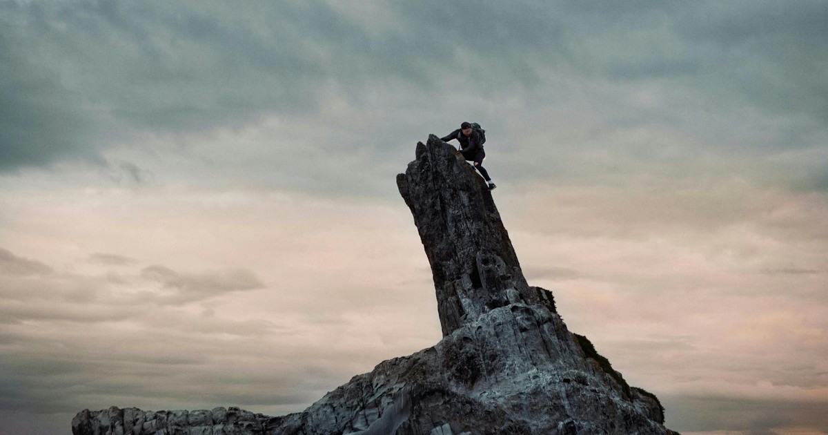 A climber scales a jagged rock formation under a dramatic cloudy sky at sunrise.