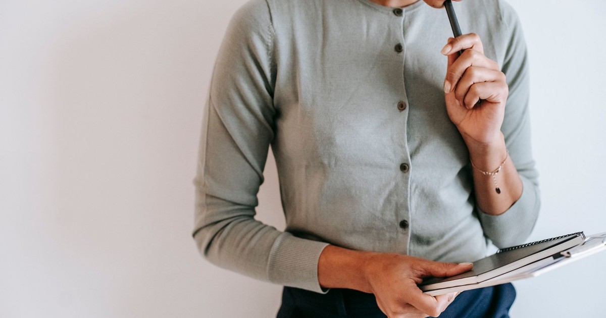 Crop unrecognizable female worker in casual clothes standing with clipboard and notebook and touching chin with pen against white wall
