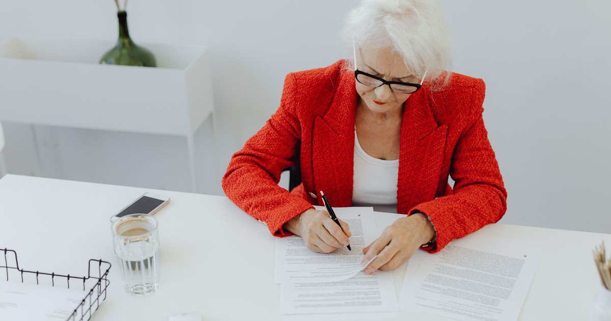 Elderly woman in red jacket writing at a tidy desk in a modern office setting.