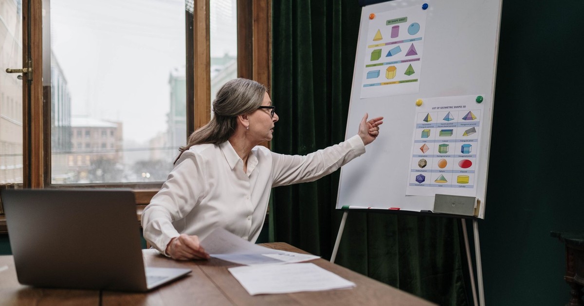 Senior woman conducting an online geometry lesson, highlighting various shapes on a chart with a laptop.