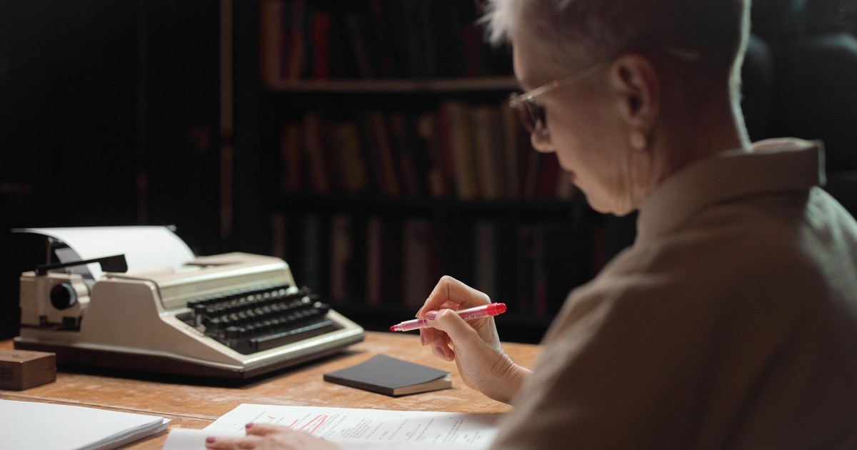 Elderly woman reviewing a manuscript with a typewriter at a wooden desk.