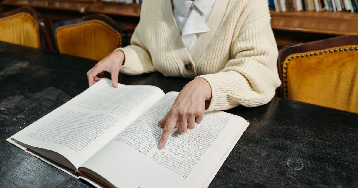 Woman pointing to text in an open book at a library's dark wooden table.