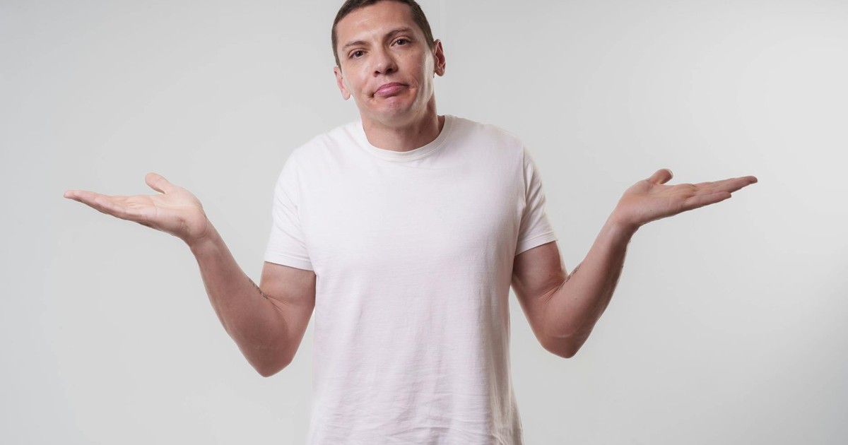 An adult man wearing a white t-shirt shrugs against a neutral background, conveying confusion or indifference.