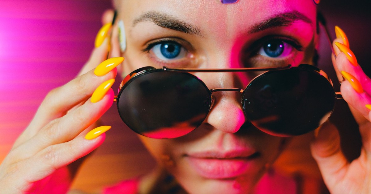 Crop bald female with multicolored stickers and bright manicure lowering stylish sunglasses and looking at camera in studio