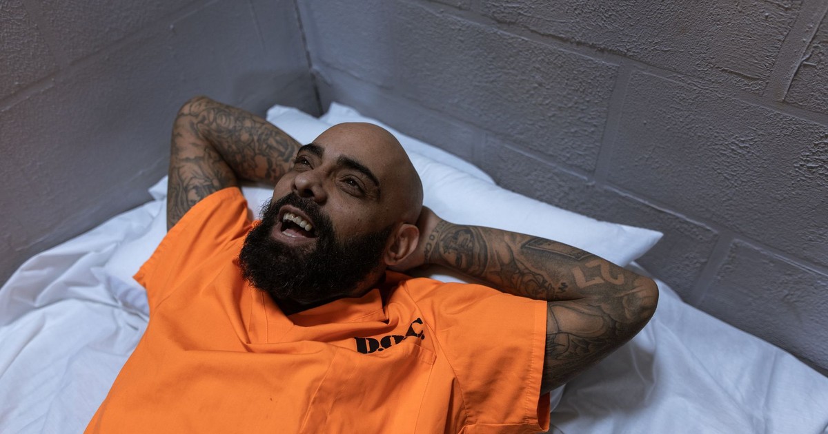 Tattooed man in orange jumpsuit smiling while lying in a prison cell.