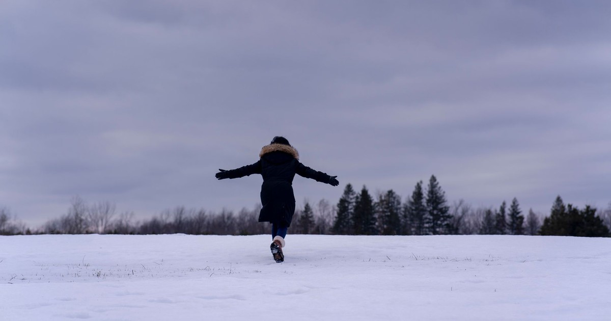 Back view of a woman in a coat running through a snowy field with arms outstretched.