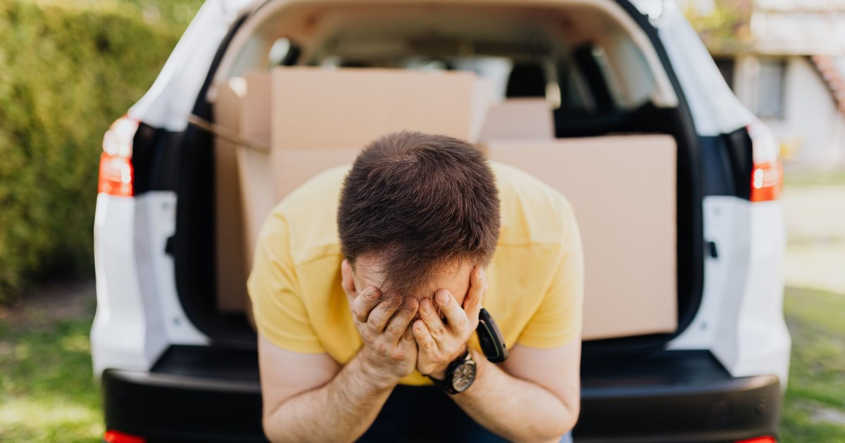 Faceless frustrated tired male wearing casual outfit sitting on car luggage boot and covering face with hands during relocation on sunny summer day