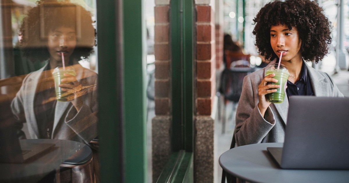 Elegant woman sipping green smoothie and working at a cafe table near a window.