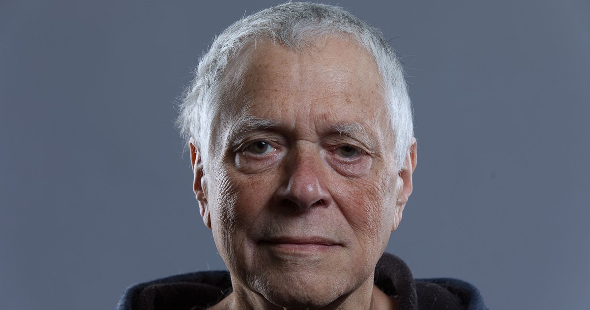 Close-up portrait of an elderly man with a neutral expression, grey background.