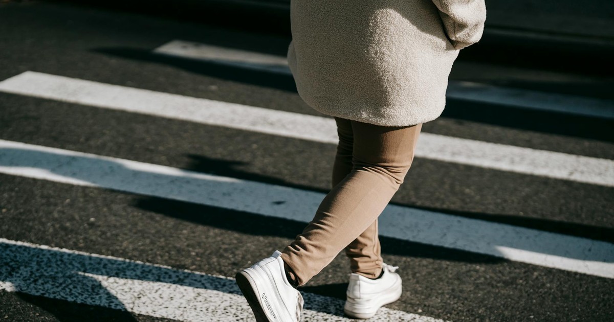 Back view crop unrecognizable female wearing casual outerwear crossing road on pedestrian crosswalk on sunny day