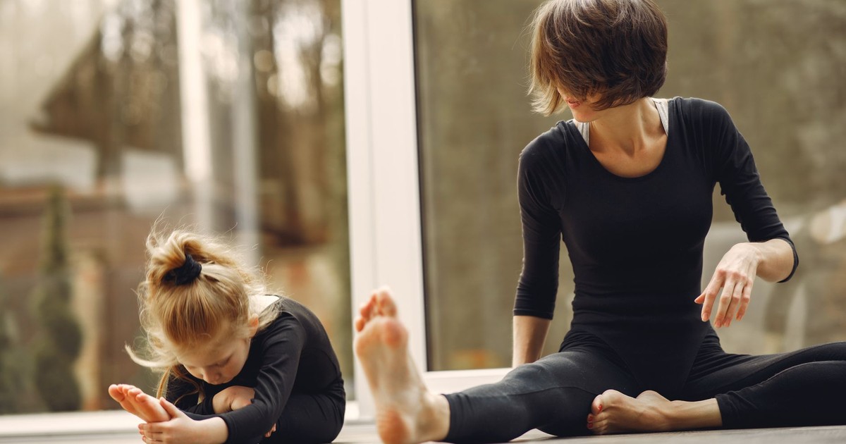 Anonymous female instructor in sportswear sitting near little blond gymnast doing tilt forward on floor near transparent door while stretching legs