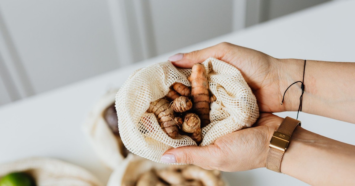Hands holding fresh turmeric in a mesh bag on a white table, highlighting natural ingredients.