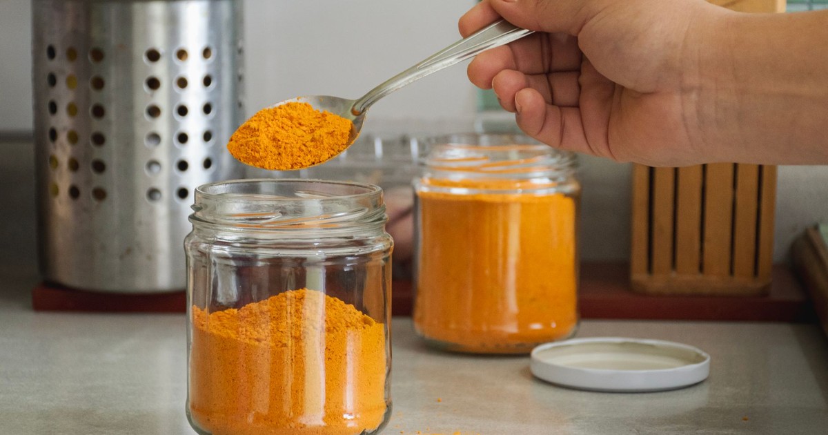 A close-up of turmeric powder being scooped into a jar in a kitchen with utensils.