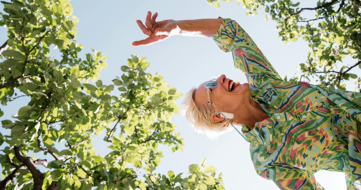 Senior woman expressing joy while dancing under green trees on a sunny summer day.