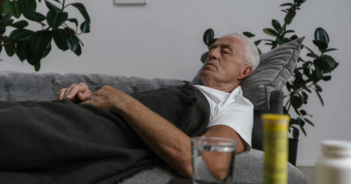 An elderly man sleeping on a couch with a blanket and glass of water nearby.