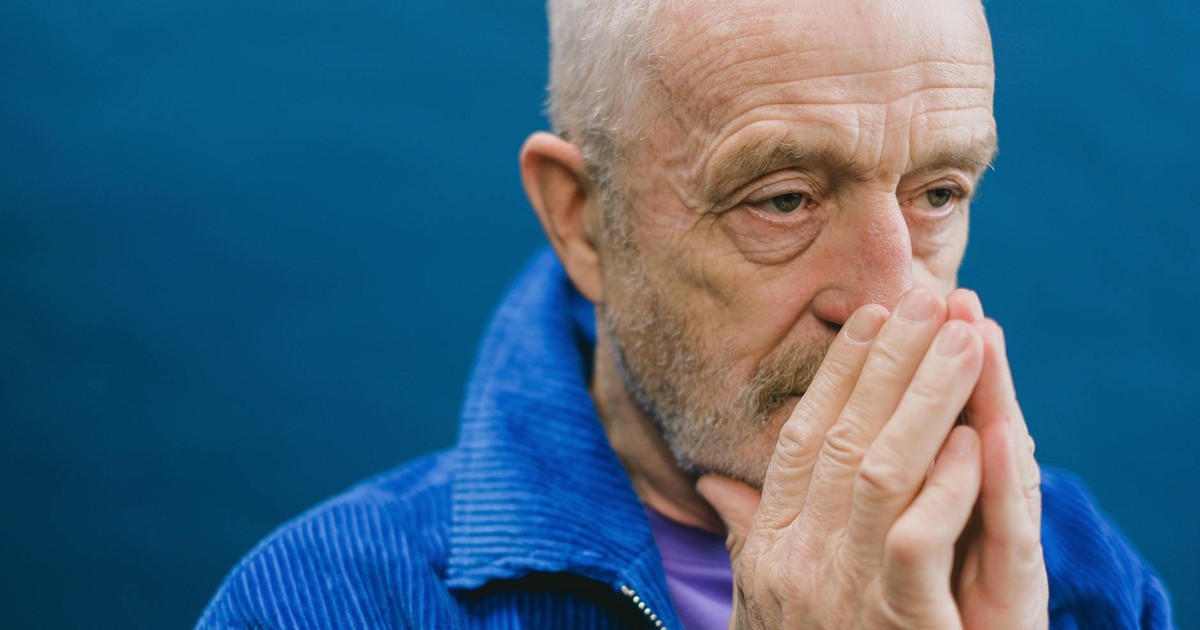Senior unshaven gray haired male in blue jacket standing against blue background with hands clasped near face while looking away