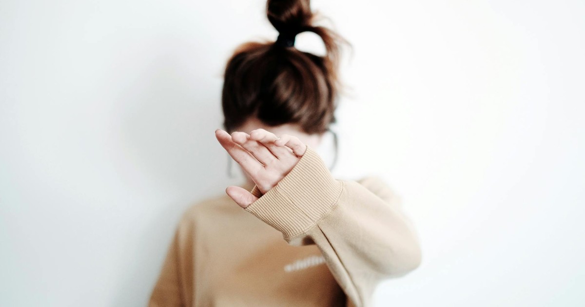 A woman in a sweatshirt raises her hand to cover her face, suggesting stop or privacy.