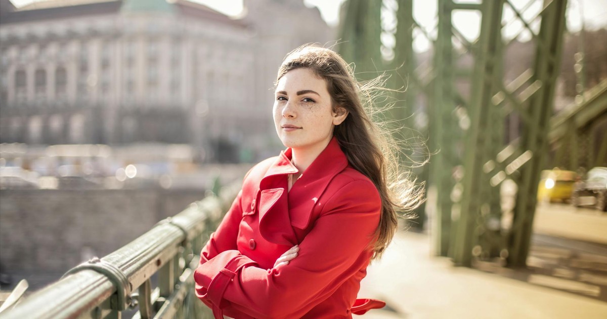 Woman in a red coat standing confidently on a bridge on a windy day.