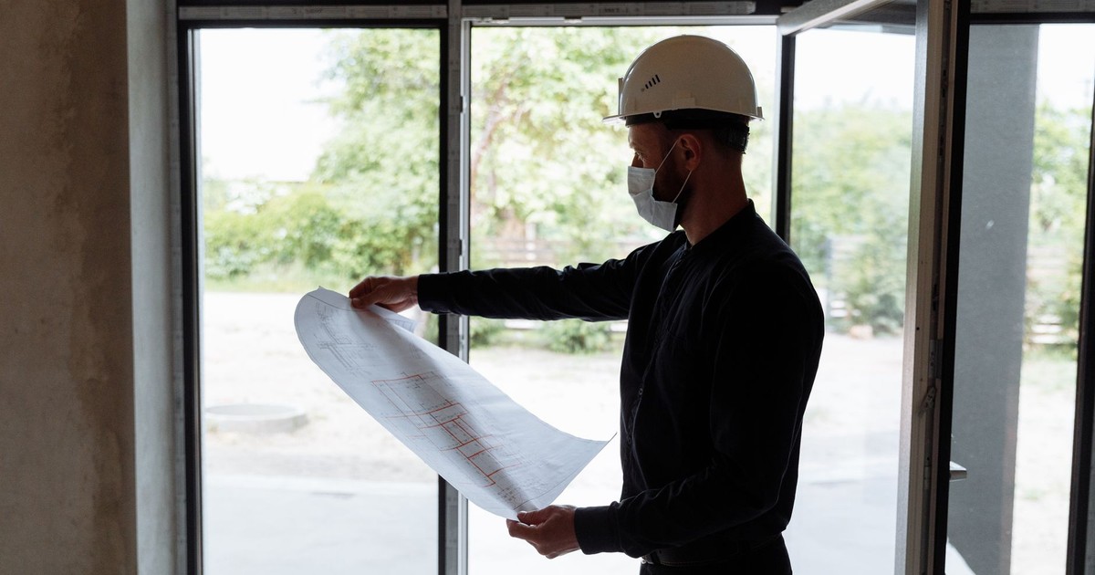 Architect reviewing floor plans indoors, wearing safety gear, focused on construction planning.