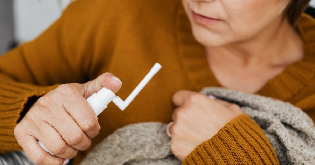 Adult woman using a mouth spray for relief indoors, wrapped in a cozy sweater.