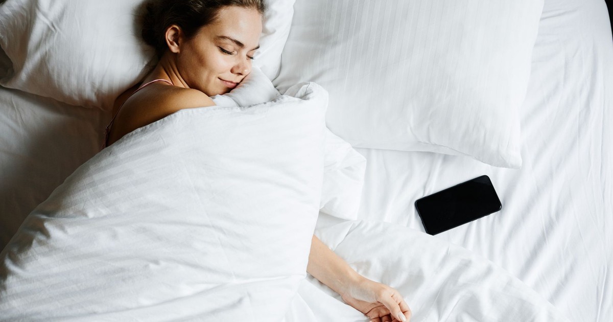 A young woman sleeps comfortably in bed with a smartphone beside her, wrapped in white sheets.