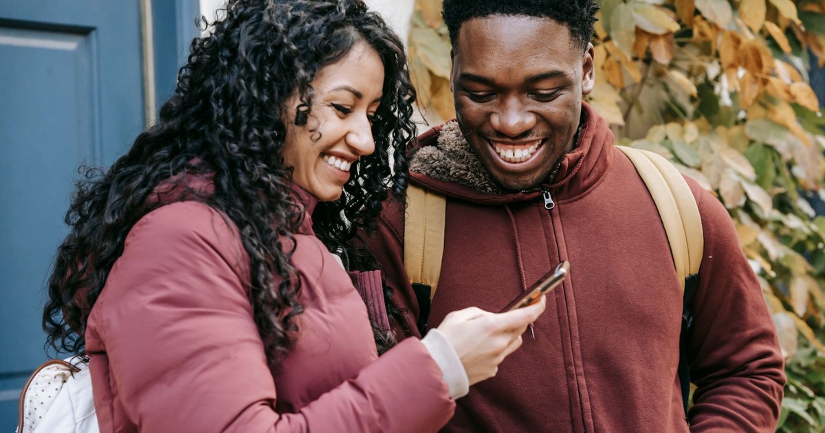 Happy multiracial couple in warm clothes smiling and using mobile phone while surfing internet on street