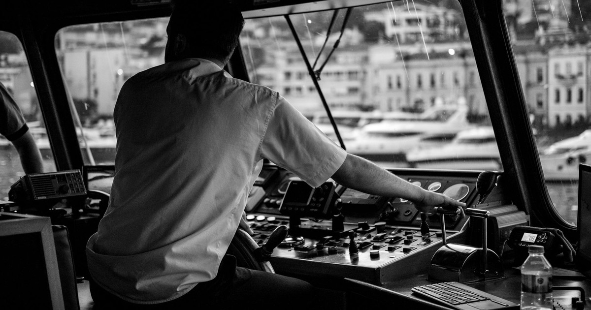 An atmospheric black and white image of a captain navigating a vessel in a busy harbor environment.