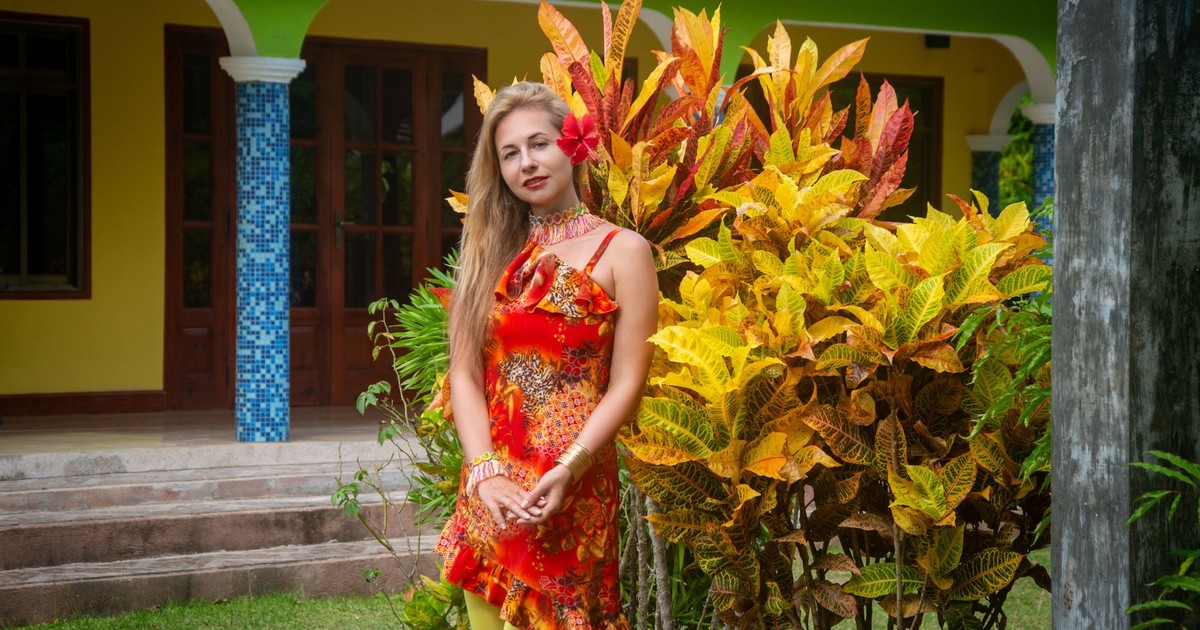 Woman in vibrant tropical dress poses by colorful croton plants in a garden.