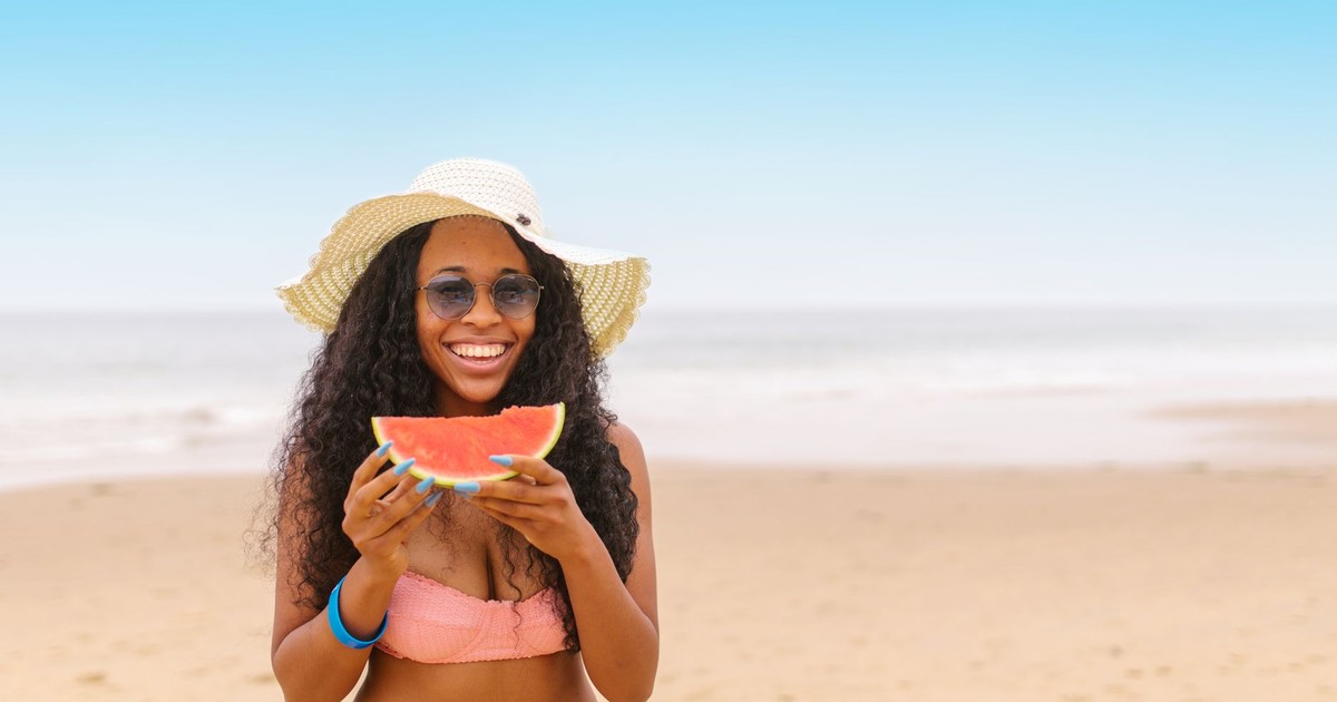 A joyful woman in a sun hat and bikini enjoys a slice of watermelon on a beach day.