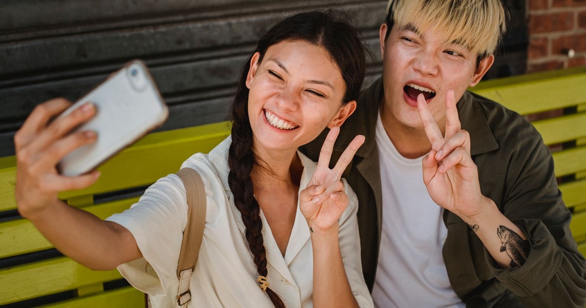Delighted young Asian couple wearing casual clothes taking selfie on modern mobile phone and showing v sign while sitting on street bench