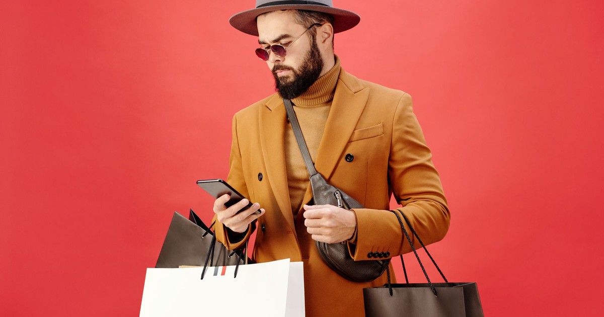 Fashionable man with shopping bags using smartphone. Stylish look with beard, hat, and sunglasses.