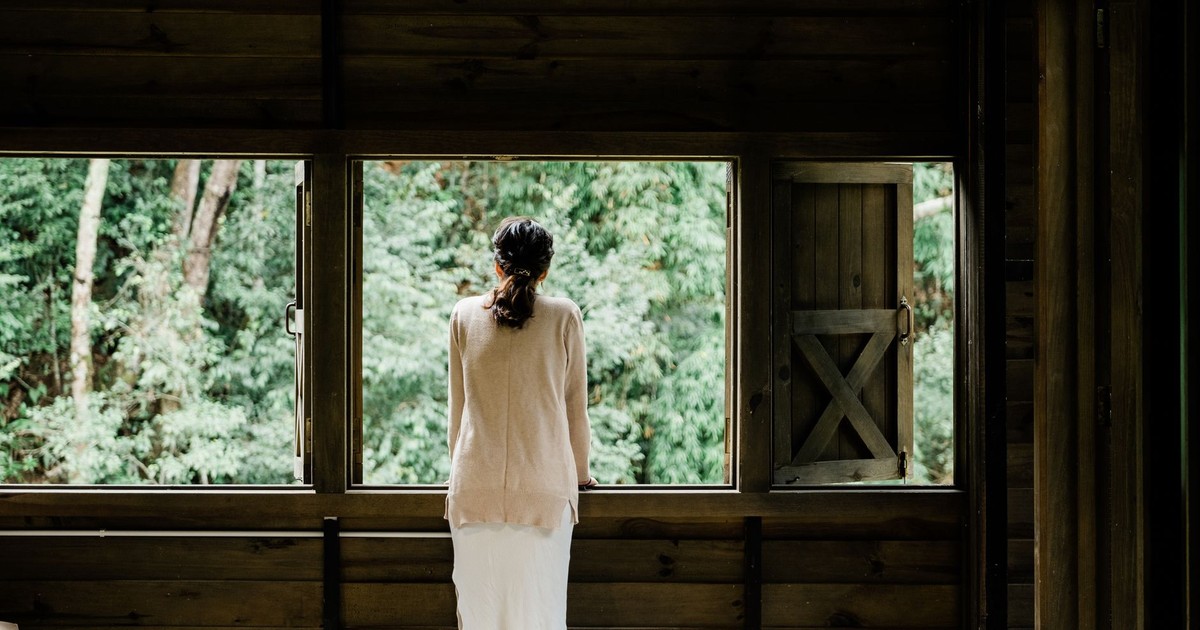 A woman stands indoors, looking out a wooden window at a lush green forest.
