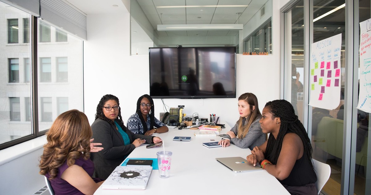 Five diverse women collaborating in a modern office meeting room, discussing projects.